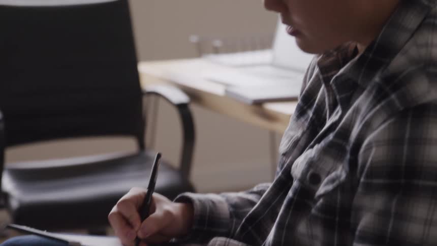 A person in a plaid shirt is seated actively writing or sketching on a notepad with a pen the scene suggests an indoor setting possibly an office with an empty chair and desk in the background