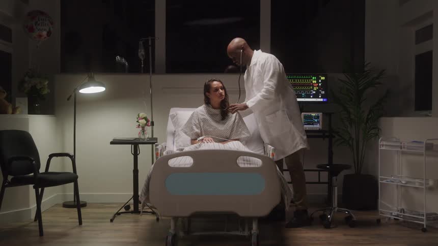 A doctor checks a female patient in a dimly lit hospital room