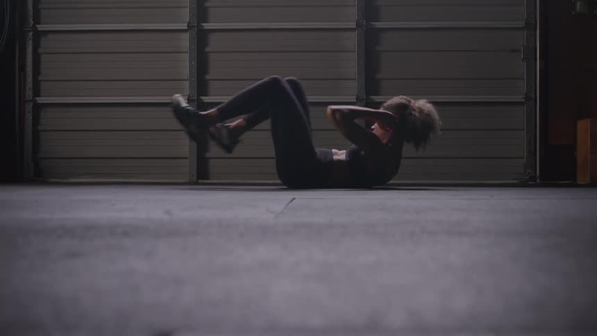A female with curly hair is performing bicycle crunches on a dark floor in what appears to be a gym or warehouse the background consists of dark ribbed garagestyle doors