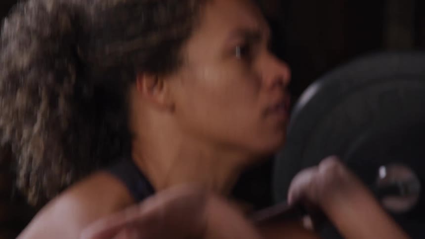 A female with curly hair is shown engaging in a workout at a gym lifting a barbell to her shoulders she is focused and determined with a glimpse of gym equipment and weights in the background