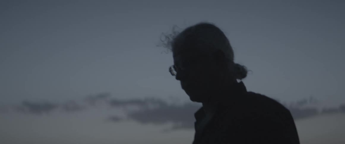 An elderly man stands outdoors in the evening
