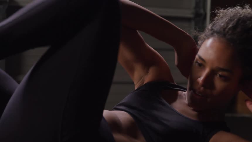 A woman with dark curly hair and a toned abdomen performs a bicycle crunch wearing a black sports bra and black leggings in a dimly lit gym