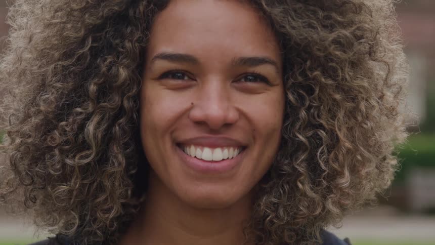 A young woman with long curly hair outdoors smiling at midday