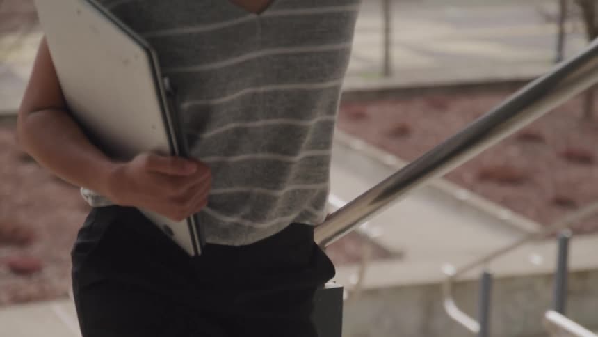 The video shows a person holding a laptop and walking up a set of concrete stairs with a metal handrail they appear to be in an outdoor urban or campus environment with buildings in the background