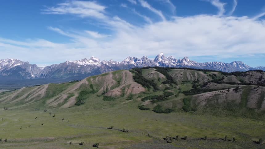 A vast landscape of grassy hills unfolds beneath a range of jagged snowcapped mountains under a vibrant blue sky