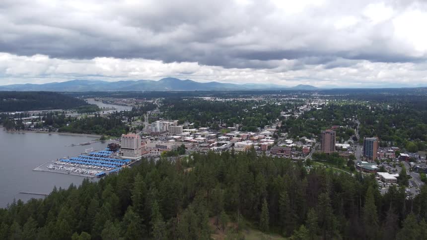 An aerial view captures a vibrant cityscape nestled beside a large body of water with dense forests in the foreground and mountains in the distant background all under a cloudy sky