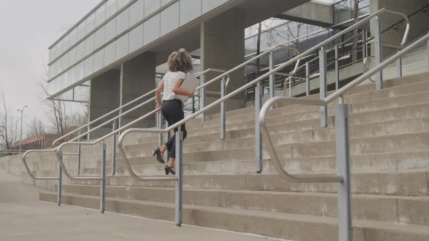 A woman in heels carrying a laptop walks briskly up outdoor concrete steps toward a modern building