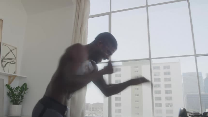 The video shows a shirtless muscular man performing what appears to be a shadow boxing or exercise routine indoors he is positioned in front of a large window with a cityscape visible in the background