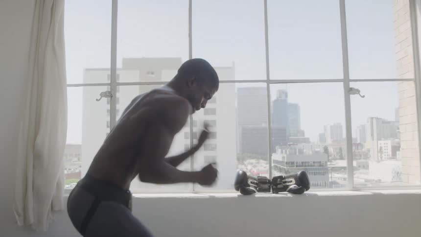 The video shows a man with dark skin wearing dark athletic shorts and no shirt standing in front of a large window he is facing to the right and making rapid punching motions with his hands as if shadow boxing
