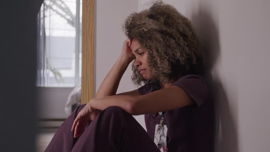 The video features a woman with curly light brown hair dressed in purple scrubs sitting on the floor with her head in her hand conveying a sense of sadness or distress the setting appears to be a hallway or a waiting area with a window