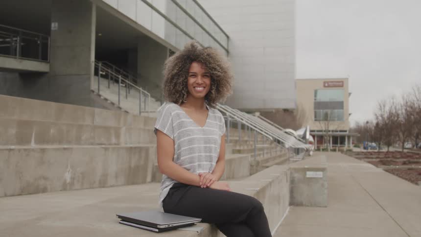 The video features a smiling woman with voluminous curly hair sitting outdoors on a concrete ledge with a laptop beside her she is wearing a striped tshirt and dark pants and a modern building with large windows and concrete stairs serves as the backd