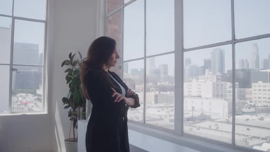A young woman looks out the window while standing with her arms crossed in an office building during daytime