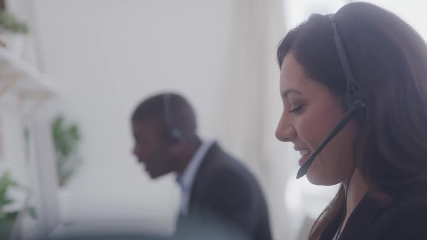 A young adult woman likely a call center agent wears a headset and looks forward with another individual sitting at a computer in the blurred background