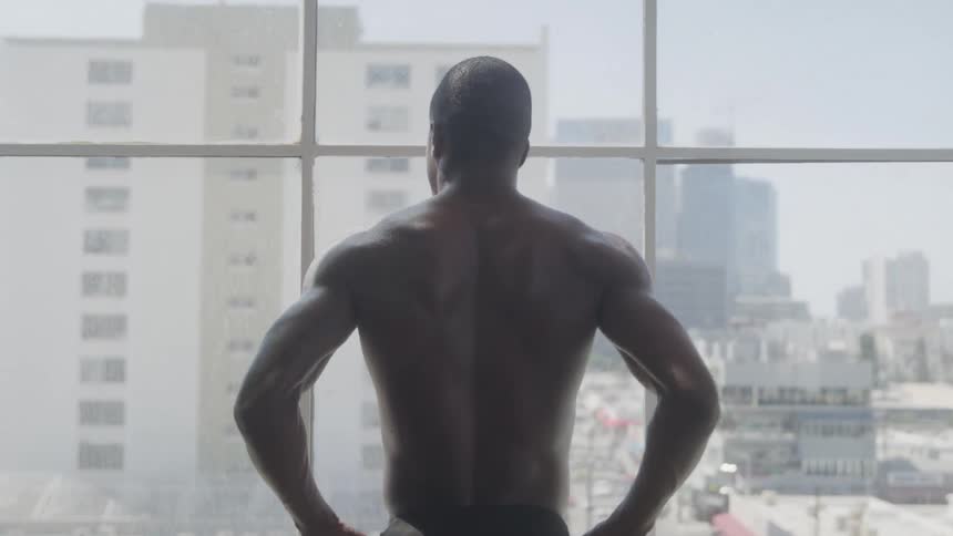 A shirtless man gazes out a large window at the city skyline