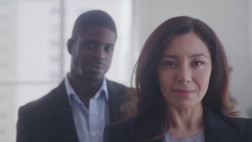 The video features a diverse duo a woman with dark hair wearing a blazer stands in the foreground initially looking away and then turning to face the camera with a subtle smile behind her a man with dark skin and a light blue collared shirt is visib