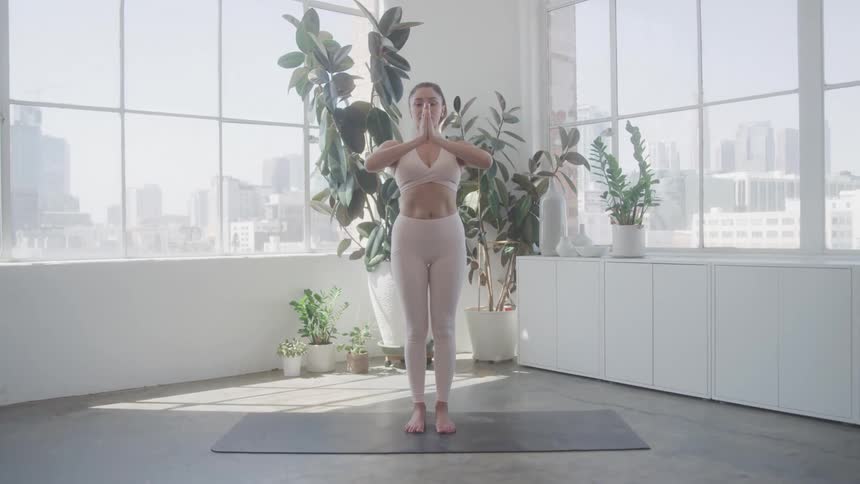 A young adult woman stands on a yoga mat in a brightly lit room with large windows overlooking a city performing a yoga pose where she raises her arms above her head and then brings her palms together at her chest