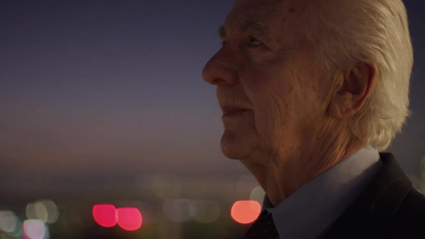 Elderly man in a suit gazes at the litup city at dusk with a serene expression