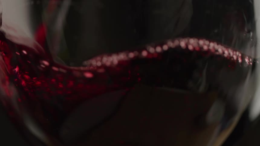 Red wine pours into a wine glass creating ripples and small bubbles as it fills the bottom part of the frame against a dark and out of focus background