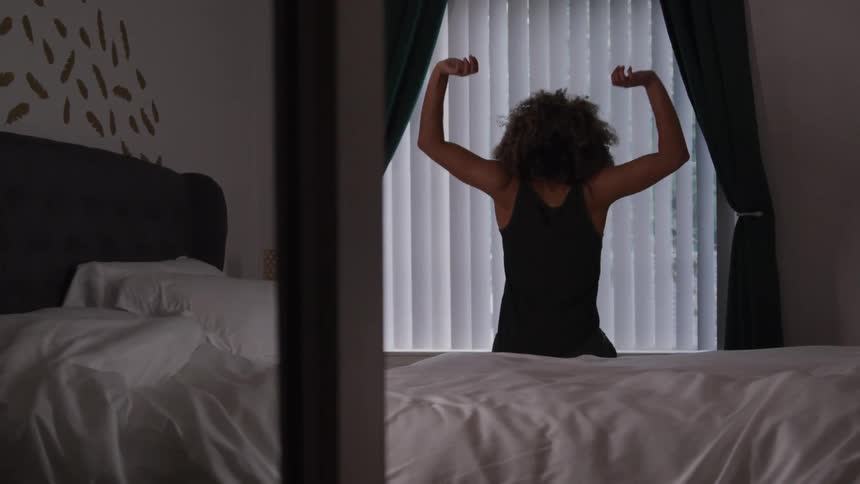 A woman sits on the edge of her bed stretching after waking up