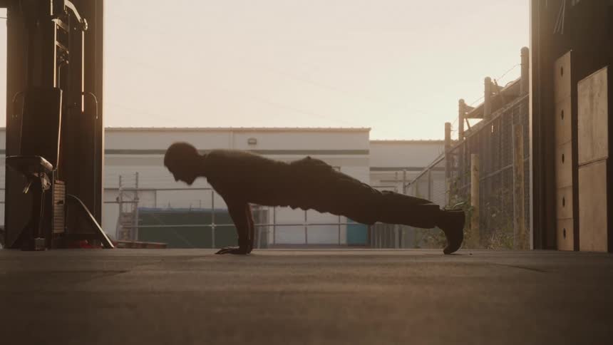 The video shows the silhouette of a person performing pushups in what appears to be a gym or warehouse setting the background is brightly lit likely by natural light coming through an open door or large window highlighting the figure in shadow