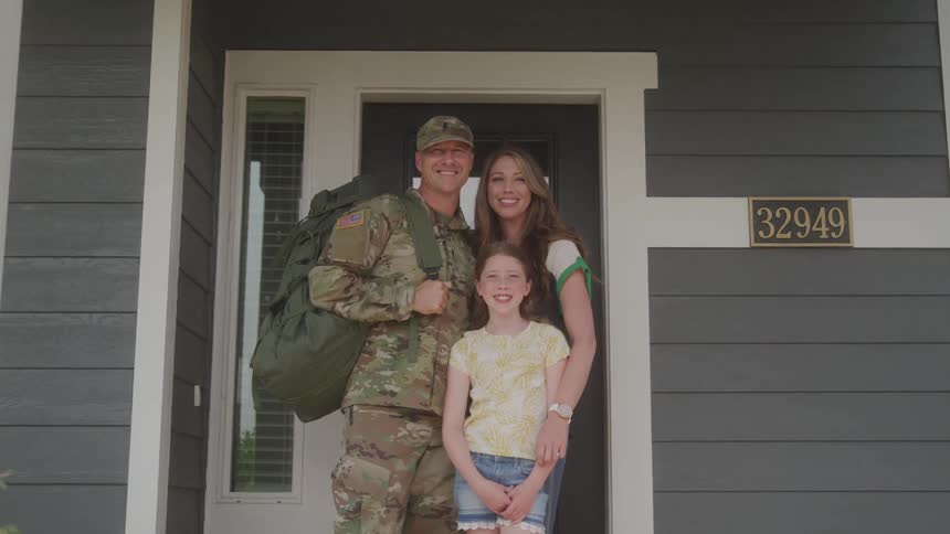 A uniformed soldier with a backpack stands smiling on the porch of a house with a numerical address embracing a woman and a young girl suggesting a joyful family reunion