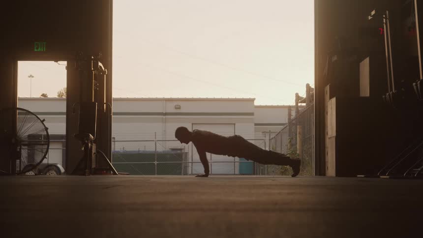 A person performs pushups silhouetted against a bright outdoor scene with buildings and a fence visible through an open doorway