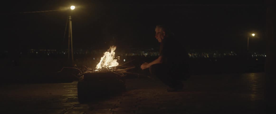 A man sits by a fire outside with city buildings in the distance