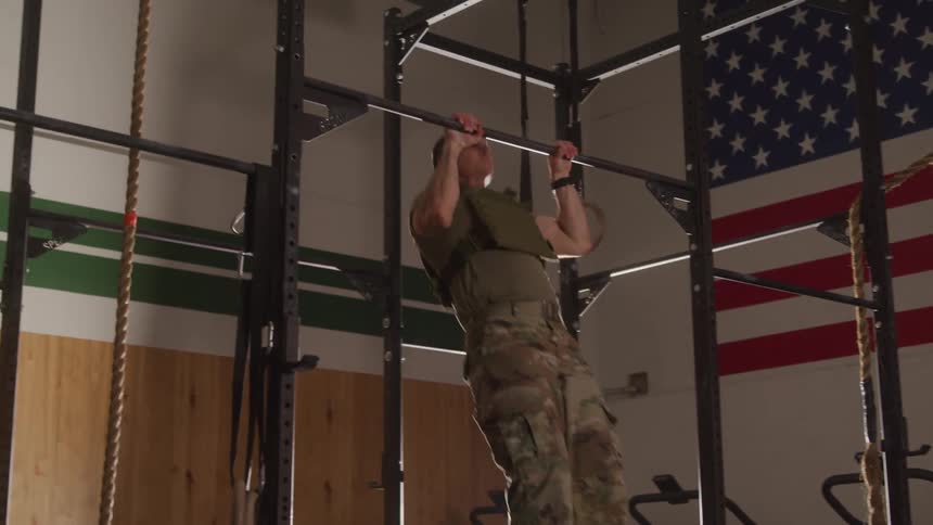 A man in military fatigues and a tactical vest performs pull ups in a gym with an american flag displayed on the wall behind him and a bright light source illuminating the scene