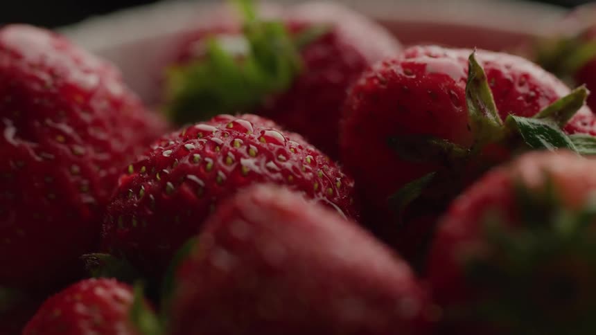 An extreme closeup with a shallow depth of field focuses on a pile of fresh vibrant red strawberries covered in tiny water droplets highlighting the rich texture of their seeds