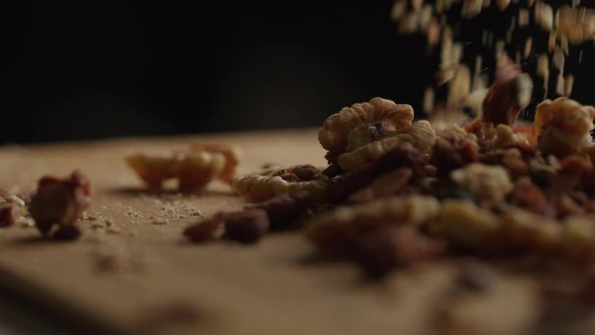 A closeup slowmotion shot shows shelled walnuts and smaller pieces tumbling onto a wooden cutting board with fine crumbs scattering as they form a textured pile against a dark background