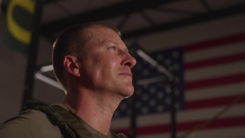 A man in military gear looks upward with a determined expression standing in front of an american flag