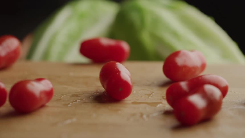 The video shows several red cherry tomatoes with water droplets on their skin being dropped onto a wet wooden surface in the blurred background there appear to be green vegetables possibly lettuce or cabbage