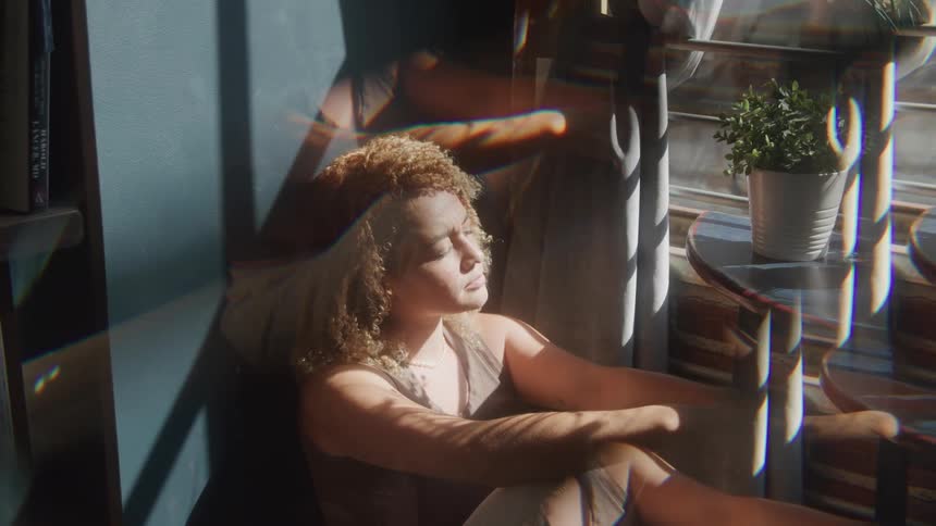 The video features a person with curly lightcolored hair sitting on the floor bathed in sunlight looking towards a window there are visual distortions possibly from light reflecting through a prism creating multiple overlapping images