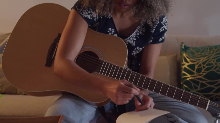 A woman with long curly hair is seated on a couch intently writing in a notebook while holding an acoustic guitar she then begins to strum the guitar appearing to play music