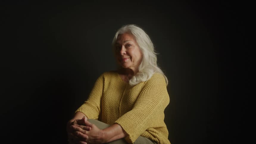 Elderly woman in a space with a dark background while standing with her legs crossed