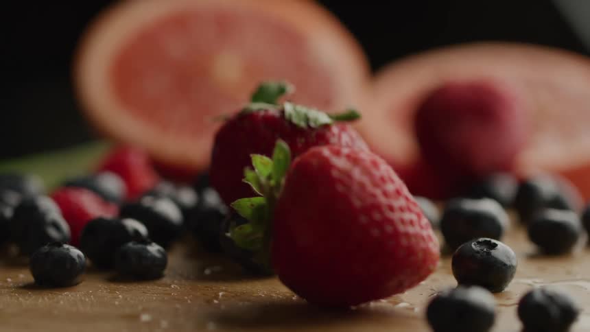 Red strawberries and dark blueberries scattered on a lightcolored surface with blurred halves of a pink grapefruit in the background