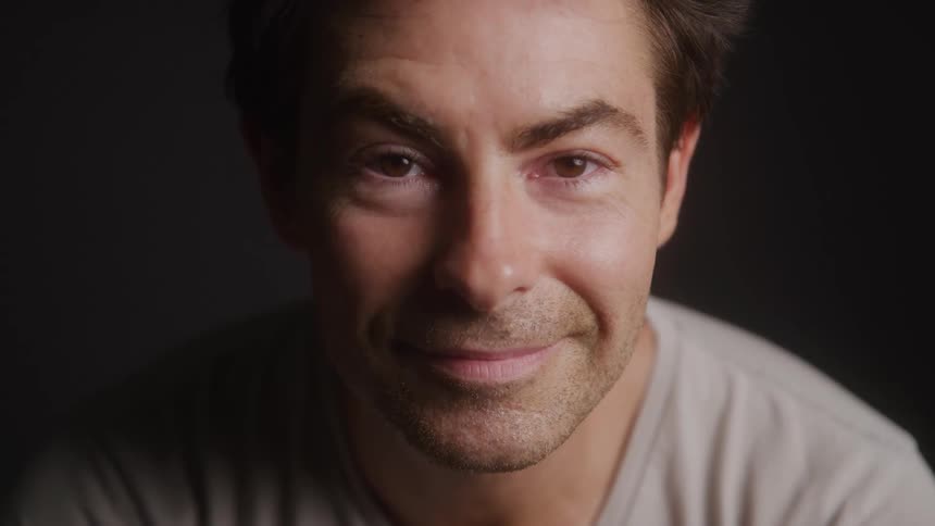 A smiling man with warm lighting on his face looking directly at the viewer against a dark background wearing a grey tshirt