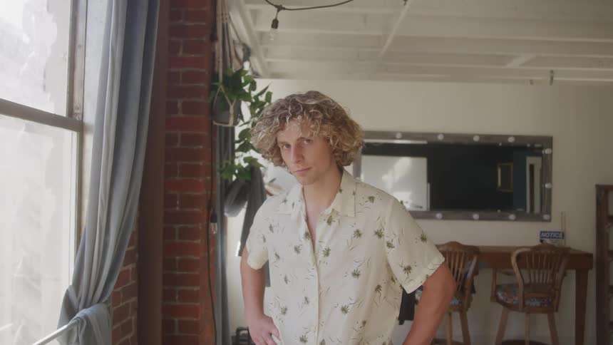 A man with curly blond hair stands in an indoor space wearing a short sleeved patterned shirt looking directly at the camera standing near a window with a grey curtain