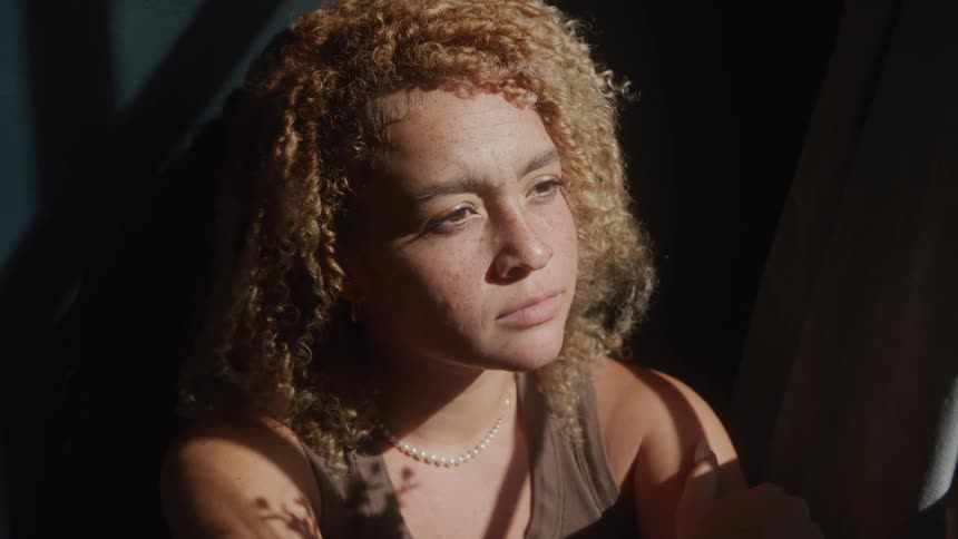 The video features a woman with curly light brown hair dressed in a brown tank top and a pearl necklace sitting indoors sunlight streams in casting shadows and highlighting her pensive or slightly troubled expression as she looks away
