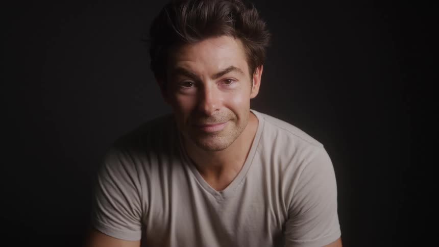 The video shows a man with dark hair looking down and then raising his head to smile slightly at the camera he is wearing a lightcolored tshirt against a dark background