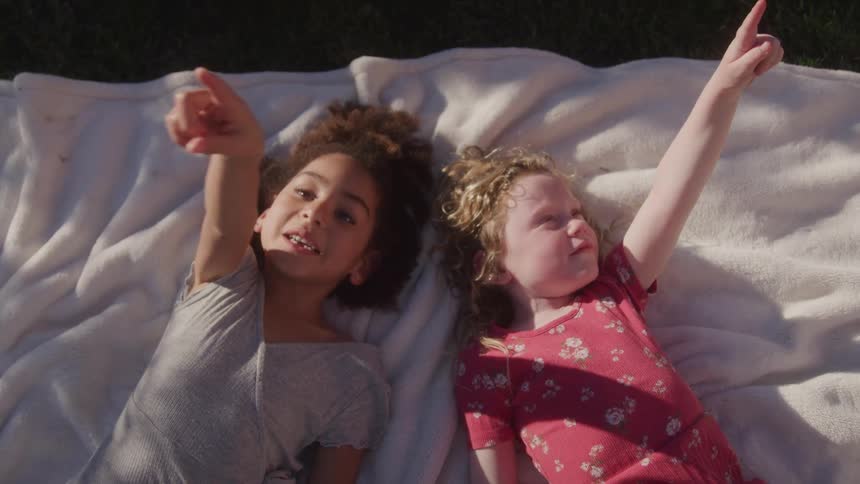 The video features two young girls one with curly dark hair and the other with curly blonde hair lying on a white blanket in a sunny outdoor setting both children are looking and pointing upwards seemingly at something in the sky