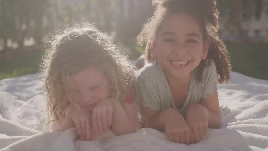 Two young girls one smiling broadly and the other with a shy expression are lying on a white blanket outdoors with sunlight creating a bright haze in the background