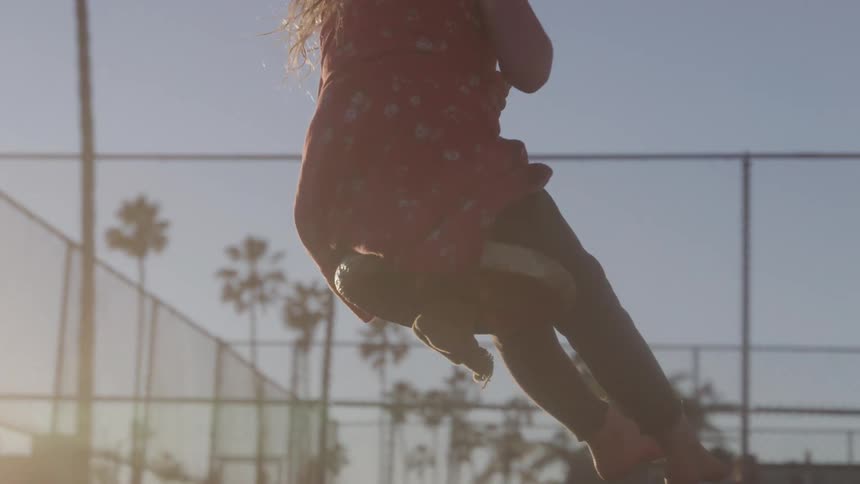 The video shows a lowangle shot of a persons legs and feet as they swing on a swing set the sun is setting in the background casting a warm glow and creating lens flare effects palm trees are visible in the distance adding to the outdoor possibly t