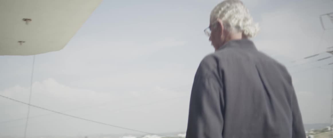 An old man walks into a house with power lines in the distance