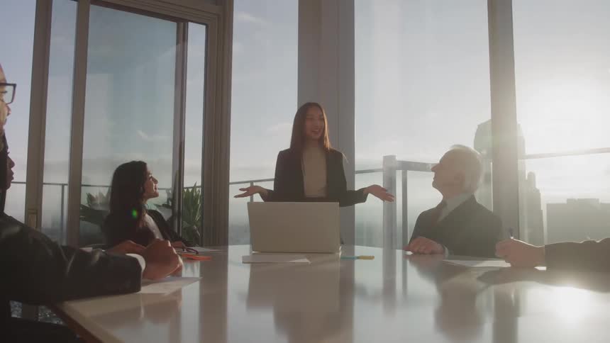 Woman standing in front of a laptop giving a presentation to some viewers in an office room