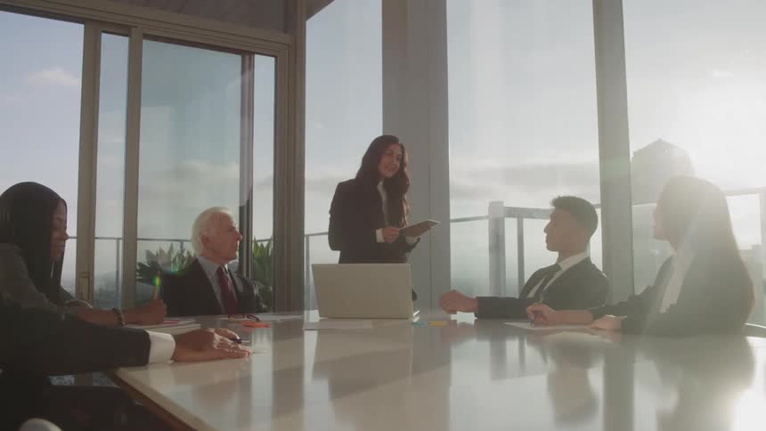 A woman standing and presenting to her colleagues in a conference room with an elderly man beside her while the others are seated and attentively listening
