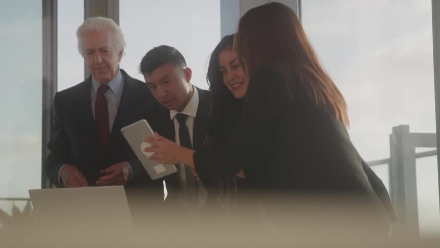 The video shows a diverse group of four professionals two men and two women gathered around a table in a bright modern office setting they are all looking at a tablet held by one of the women seemingly discussing its content or collaborating on a tas