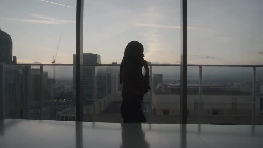 A woman silhouetted against a panoramic skyline paces indoors on a phone call as the sun sets beyond the highrise windows
