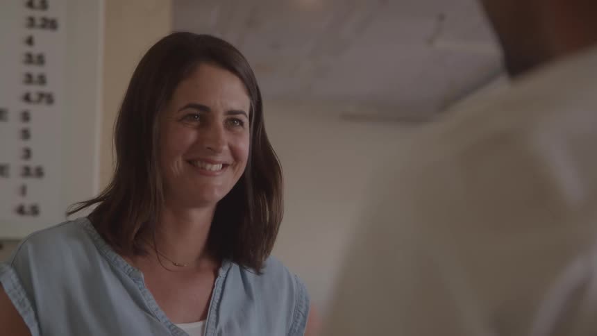 A woman with shoulderlength dark hair is smiling and looking at someone offcamera she is wearing a light blue top and a delicate necklace in the background a menu board with numbers is visible
