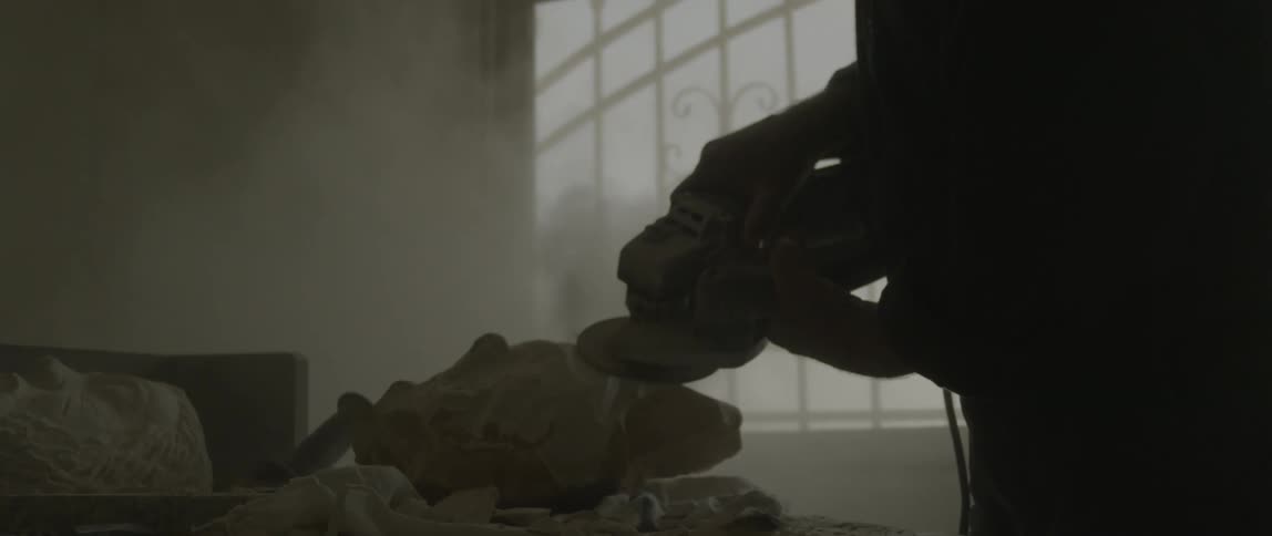 A man sits by a window sanding some stone